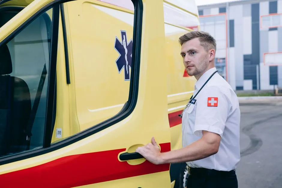A young male paramedic in uniform opens the door of a yellow ambulance outdoors.